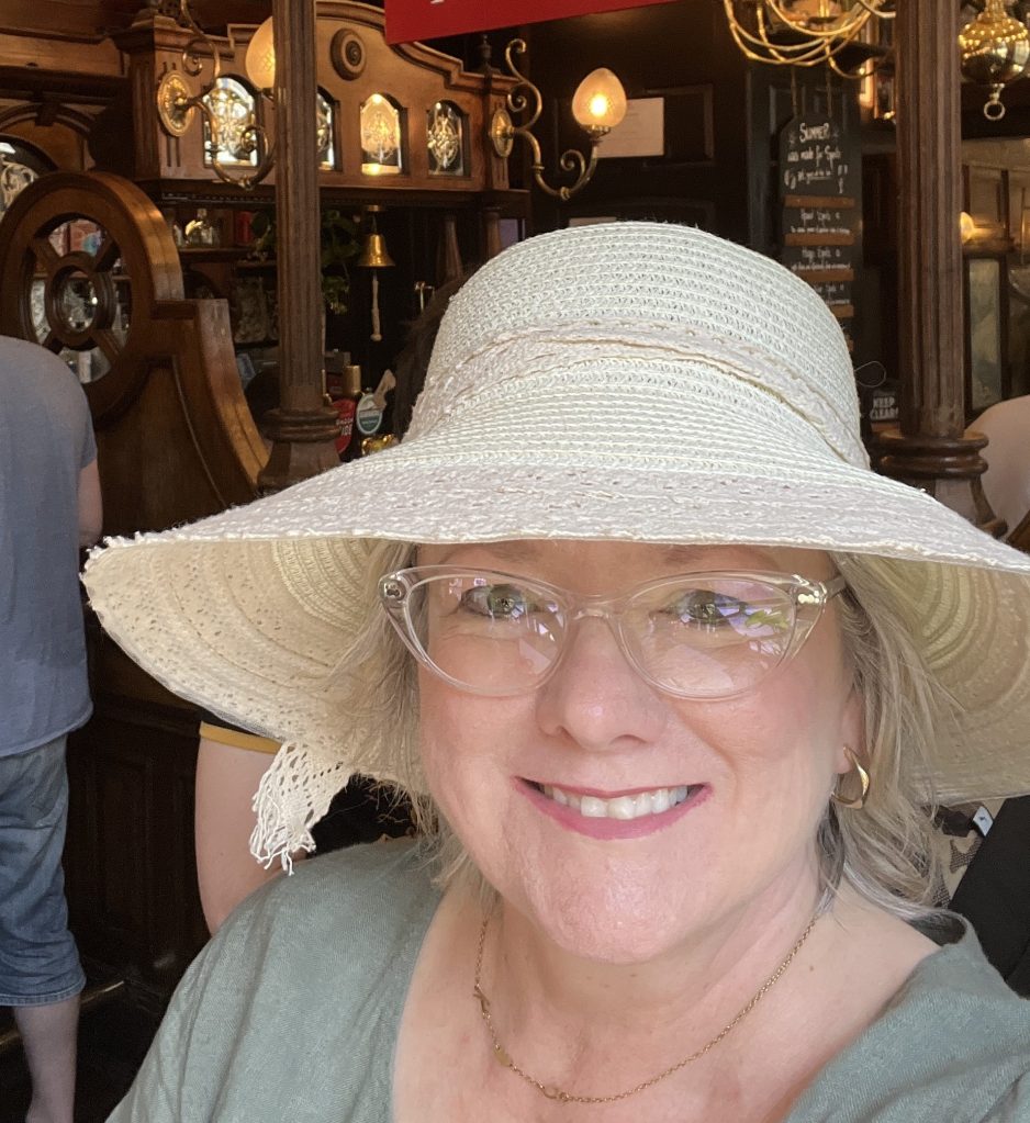 A woman smiling while wearing a wide-brimmed straw hat and clear glasses, inside a cozy bar with wooden decor in the background.